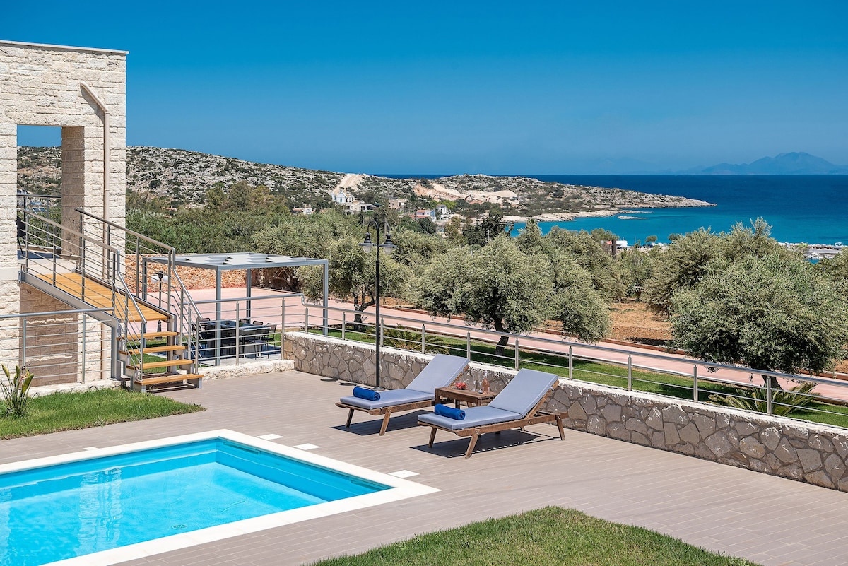 A private pool is surrounded by a stone terrace, featuring comfortable loungers under a clear blue sky. Verdant landscaping and olive trees enhance the outdoor space, while a scenic view of the coastline and hills is visible in the background.