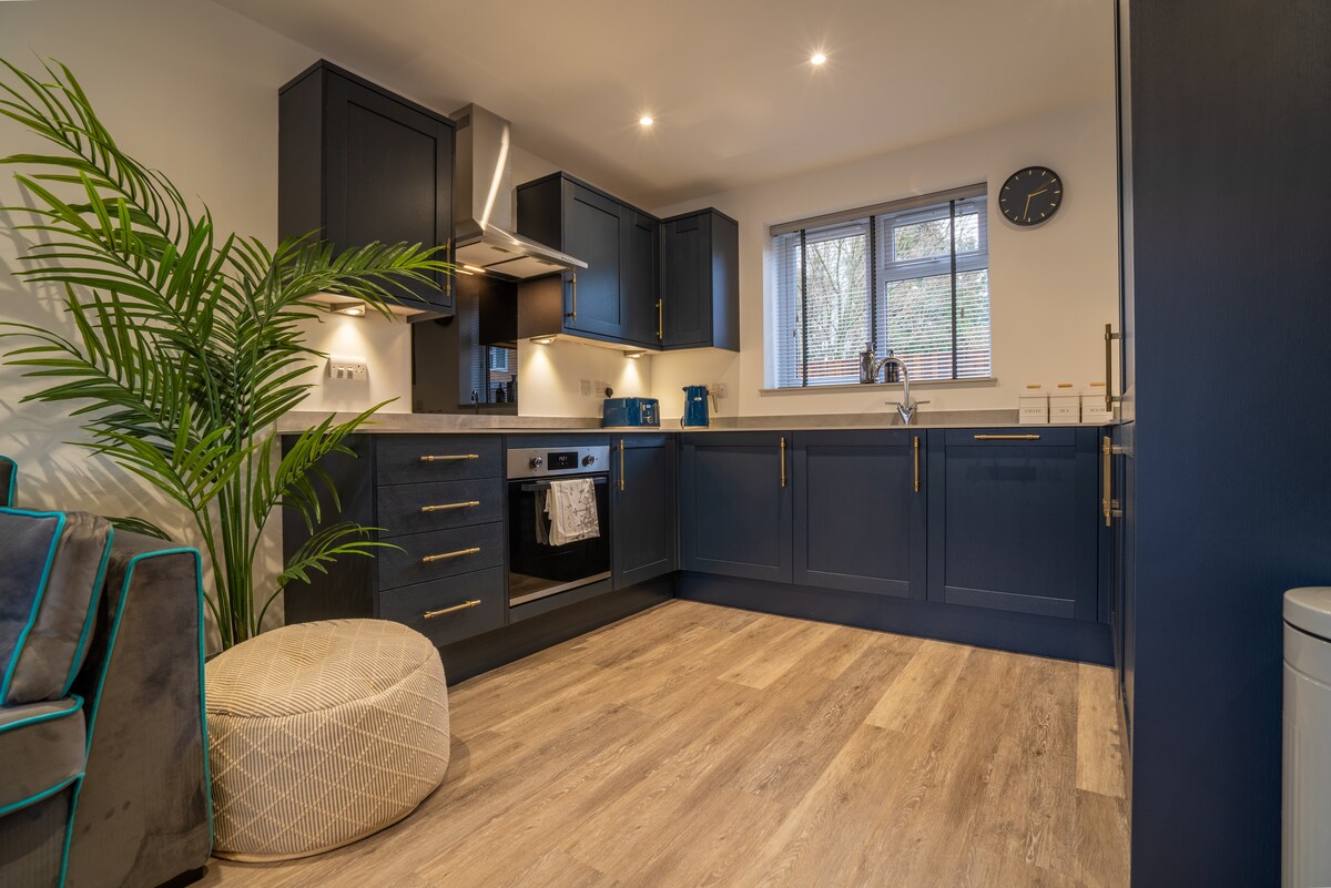 The kitchen area is fitted with dark cabinetry and modern appliances, including an oven and refrigerator. A window allows natural light to illuminate the space. A green plant adds a touch of nature, and a light-colored pouf offers additional seating.