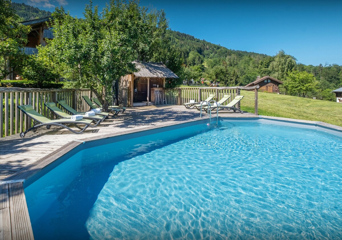 A crystal-clear swimming pool is surrounded by wooden decking and features several loungers arranged for relaxation. Lush greenery and trees border the area, providing a serene backdrop. A traditional wooden structure is visible nearby, along with expansive views of the landscape.