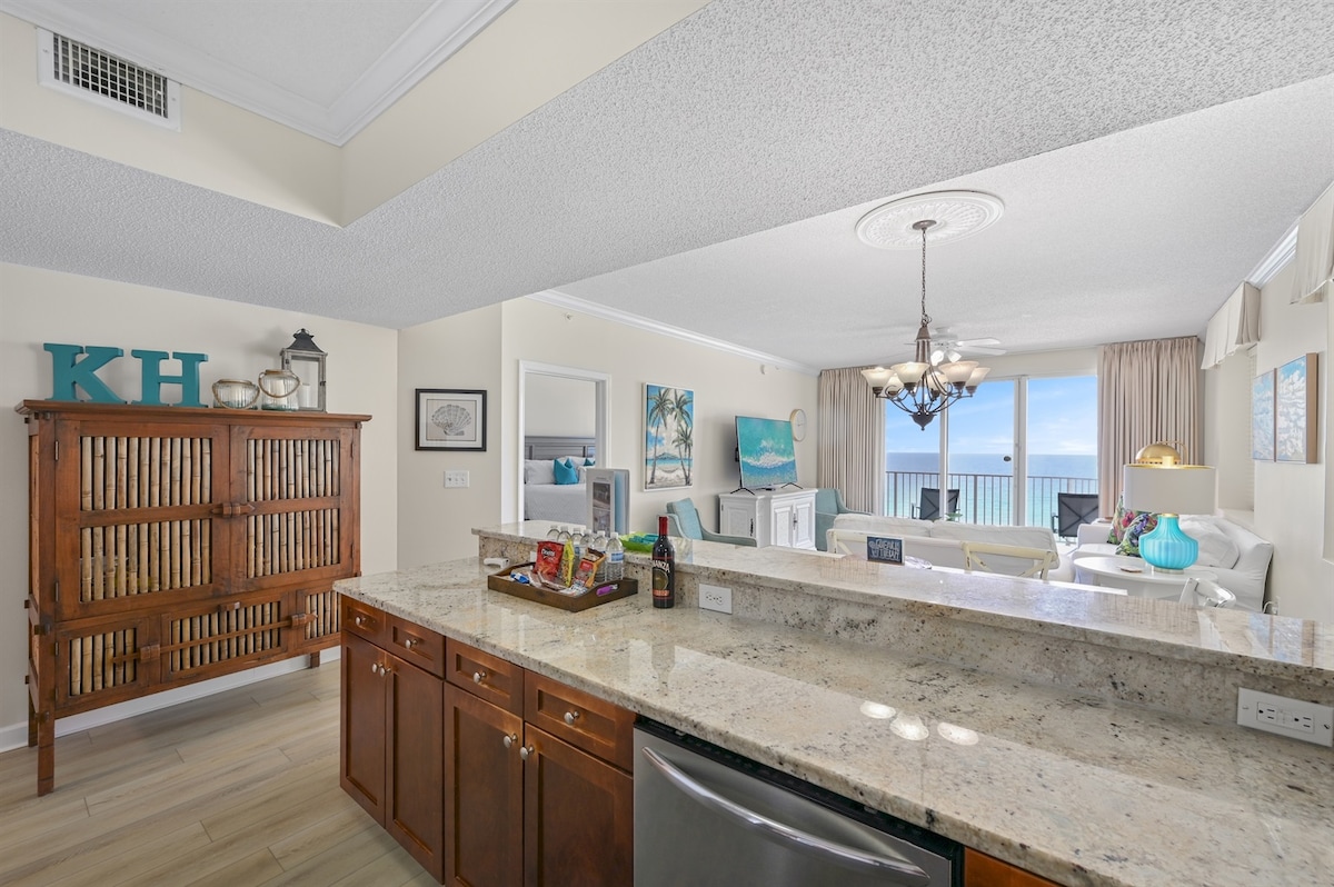 A modern kitchen area is featured with dark wood cabinetry and a spacious granite countertop. The open layout allows views of the light-filled living area, decorated with coastal art and access to a balcony overlooking the Gulf. Natural light enhances the inviting ambiance.