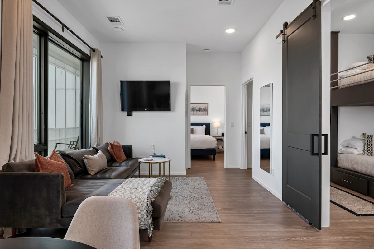 The modern living area includes a dark sofa with orange and gray pillows, a flat-screen TV mounted on a white wall, and a round table for dining. A sliding black barn door leads to a bedroom with two twin bunk beds, while natural light filters through large windows with soft curtains.