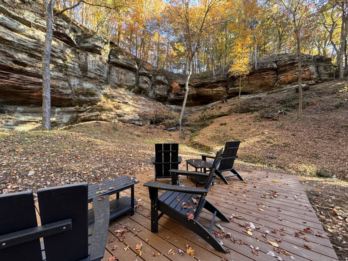 A spacious outdoor seating area features several black chairs arranged on a wooden deck. The surrounding landscape showcases autumn foliage on rocky terrain, with a cliff in the background. Sunlight filters through the trees, highlighting the peaceful retreat.