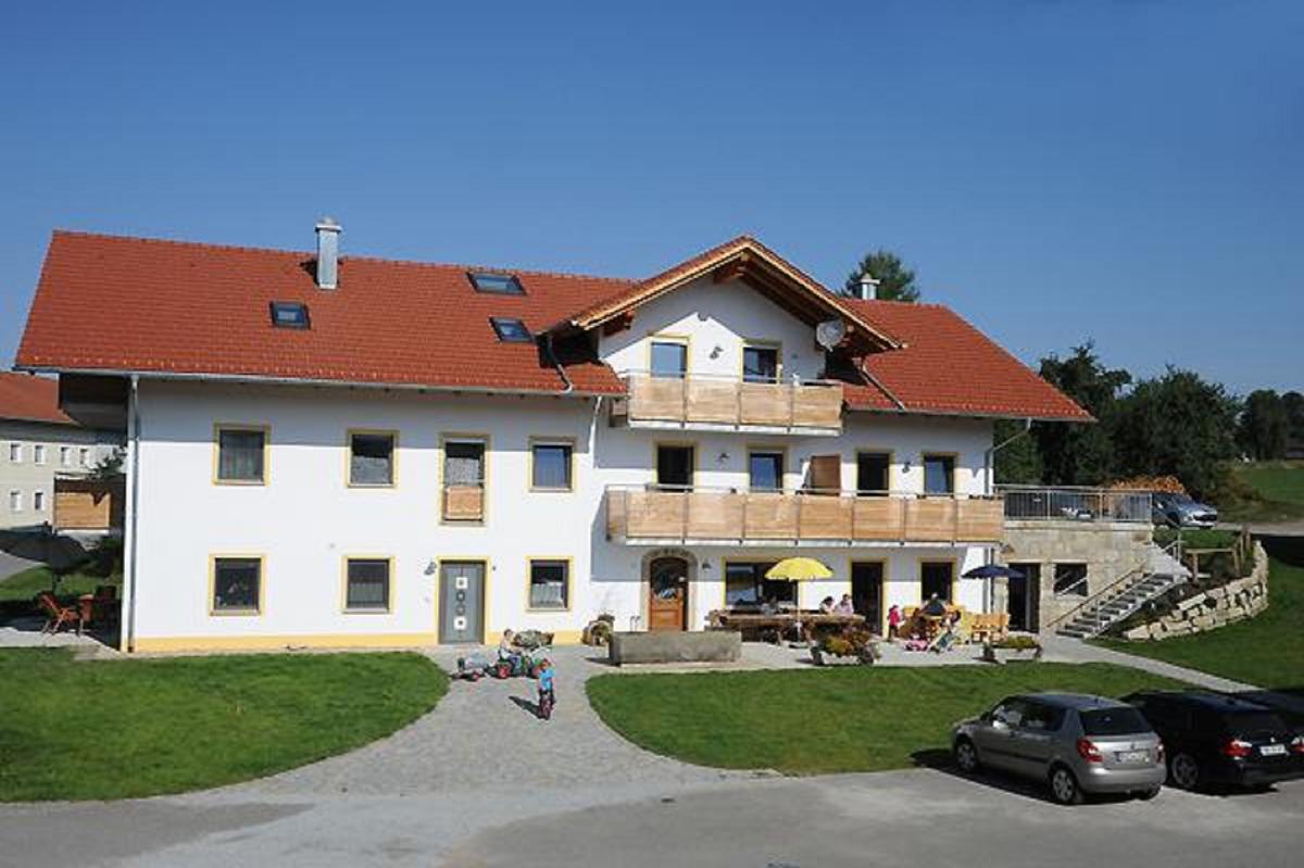 A spacious two-story building is set against a clear blue sky. The exterior features a white facade with wooden balconies and a red roof. A well-maintained lawn leads to the entrance, where guests can be seen enjoying the outdoor seating area.