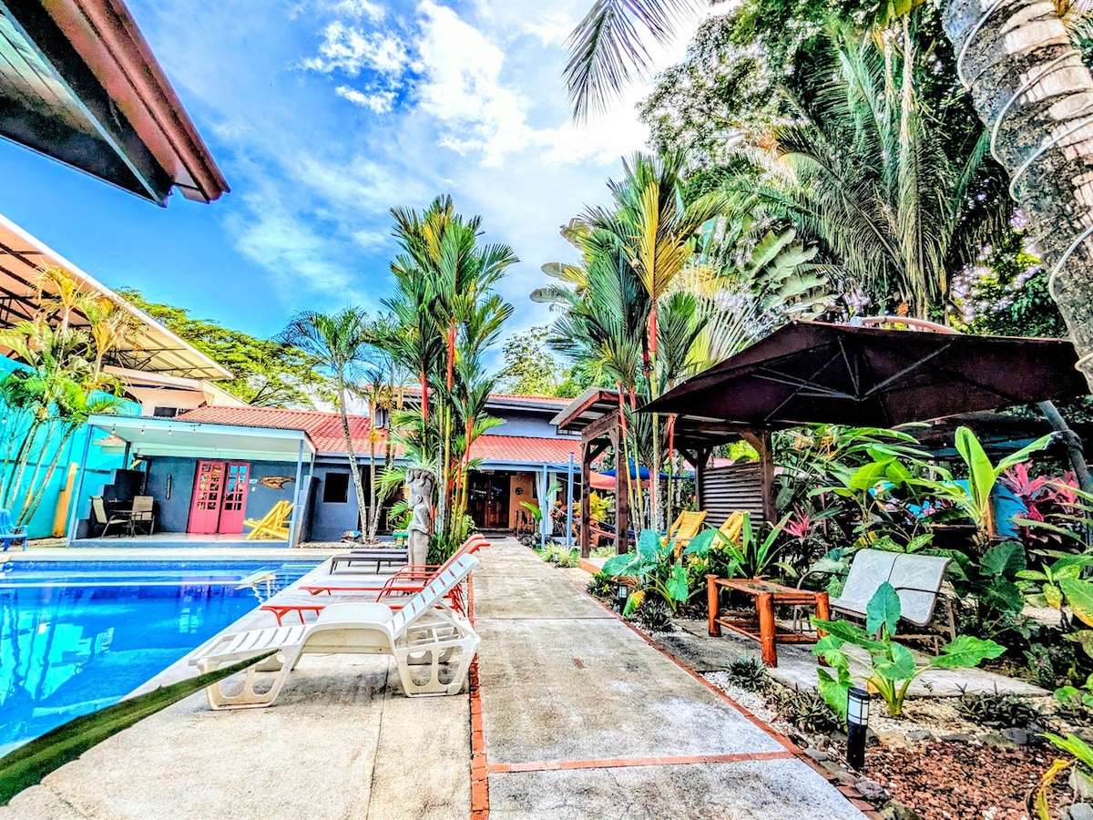 A vibrant outdoor area surrounds a sparkling pool, flanked by lounge chairs and tropical landscaping. A pathway leads toward colorful cabinas, with lush palm trees and flowering plants adding natural beauty to the setting. The inviting atmosphere is enhanced by shaded seating arrangements.