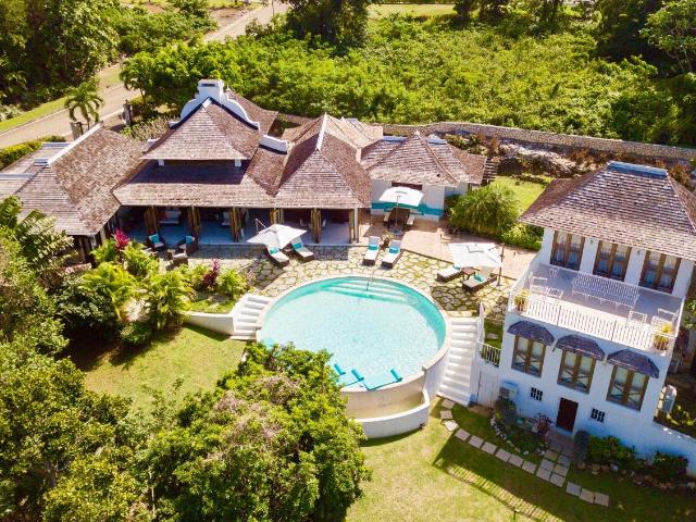 An aerial view of Half Hill villa features a circular swimming pool surrounded by well-maintained gardens. Two distinct buildings can be seen, with the main villa presenting a traditional Caribbean style and the cottage beside it. Sun loungers are positioned invitingly around the pool.