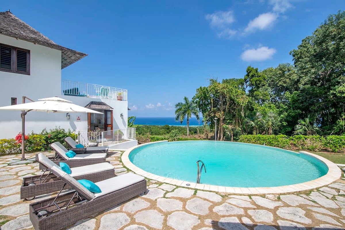 An inviting circular infinity pool is surrounded by stone tiles, with comfortable lounge chairs positioned nearby. Lush greenery and palm trees frame the backdrop, providing a serene atmosphere. A glimpse of the Caribbean Sea can be seen in the distance under a clear blue sky.