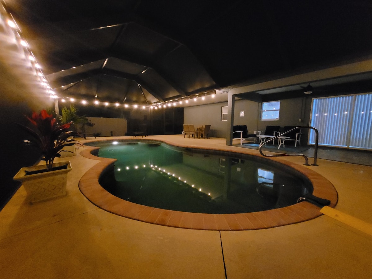 The screened-in pool area features a curved pool surrounded by a tiled deck. Soft lighting is suspended overhead, creating a relaxed ambiance. Nearby, a seating area is visible, complemented by potted plants to enhance the inviting atmosphere.