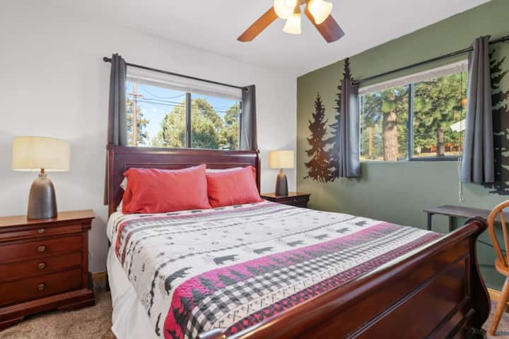 Светла спалня с весели спални принадлежности и големи прозорци, които внасят естествена светлина.