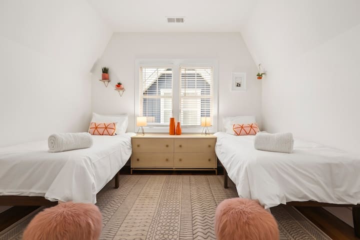 3rd Floor Twin Bedroom-Bright and inviting, this charming attic room offers twin beds perfect for a peaceful night's sleep. Adorned with pops of orange decor, the space features a central dresser and window to let in natural light.