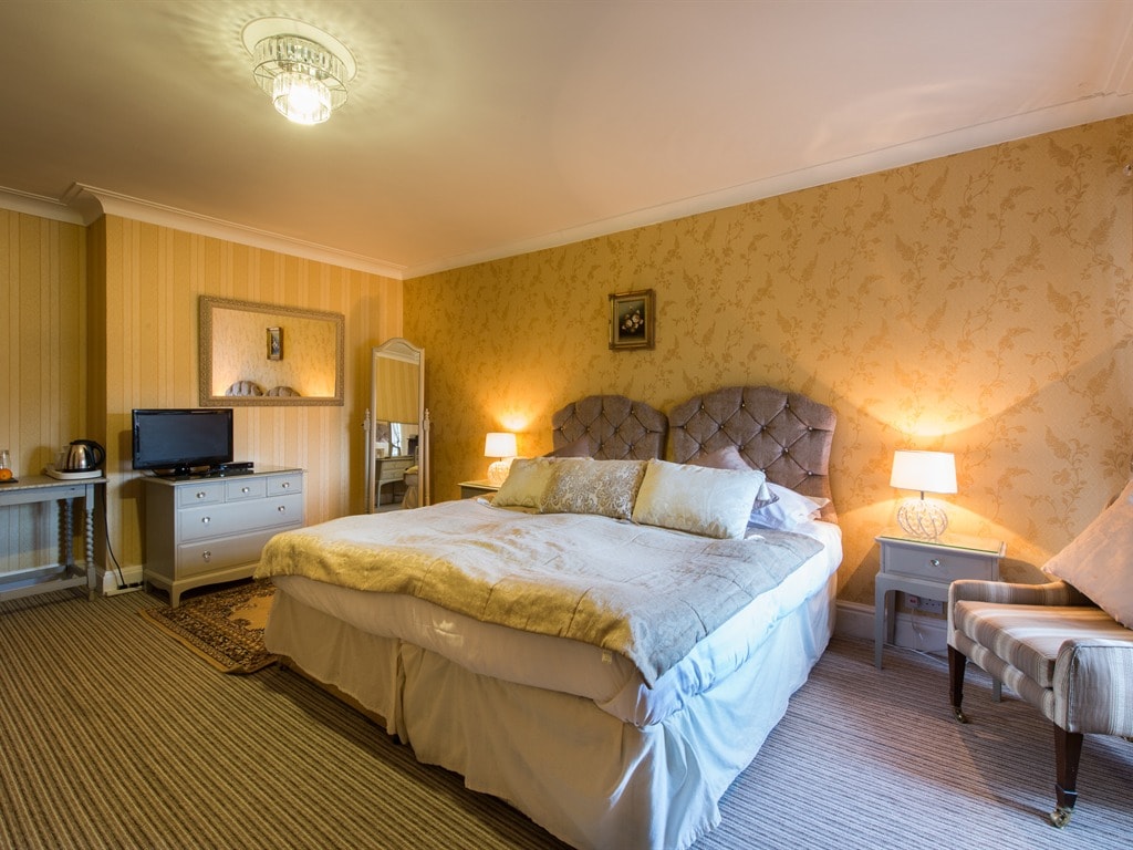 A spacious room features a large bed with a light-colored coverlet and plush pillows. Soft lighting from bedside lamps creates a calm ambiance. A chic mirror and a flat-screen television are visible against a backdrop of patterned wallpaper, while a cozy armchair adds to the comfort.