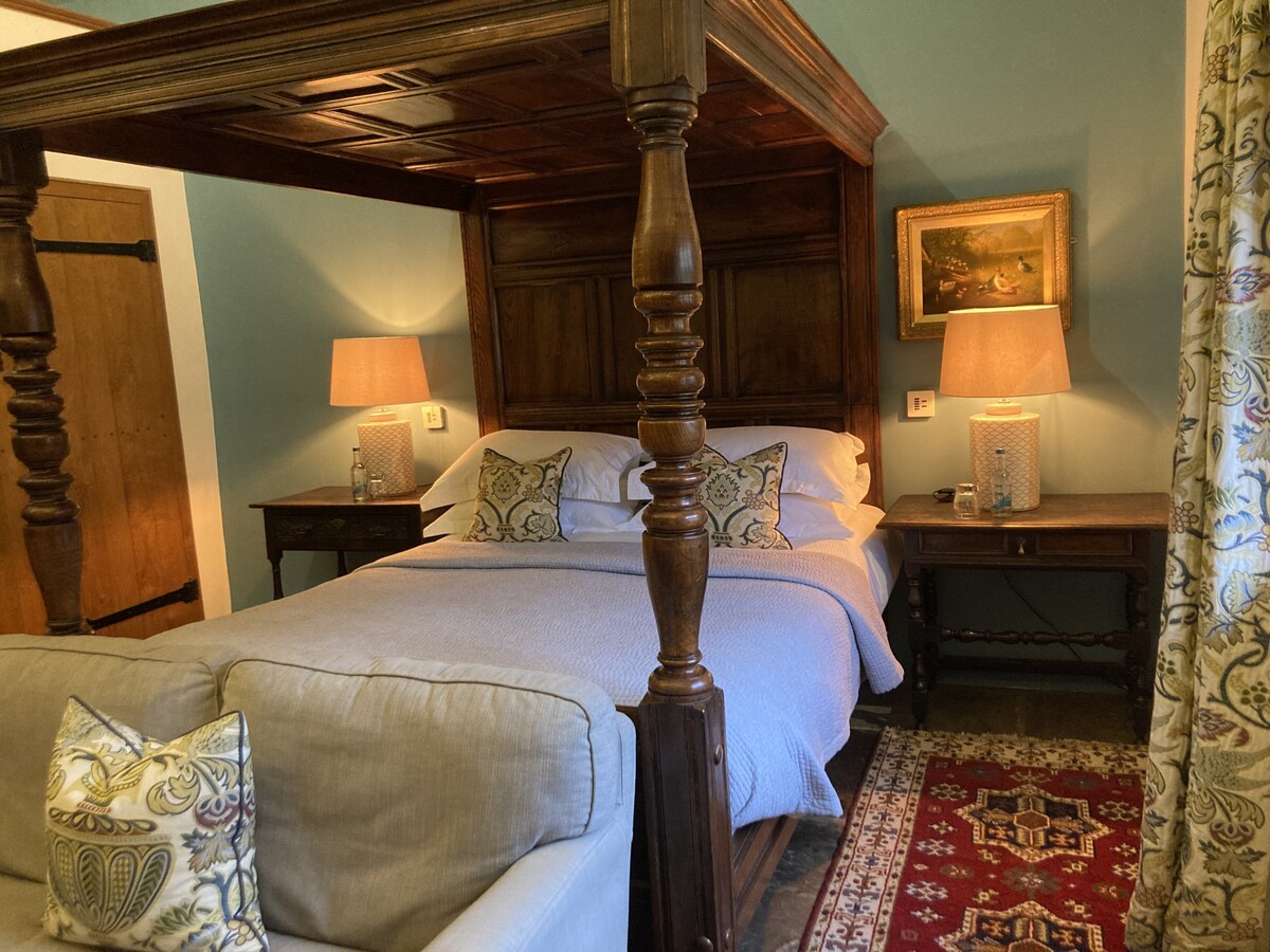 A four-poster bed is positioned centrally, flanked by two wooden nightstands with lamps providing soft illumination. A sofa with decorative cushions sits nearby, creating a cozy sitting area. An ornate rug is laid on the floor, and artwork can be seen on the wall.
