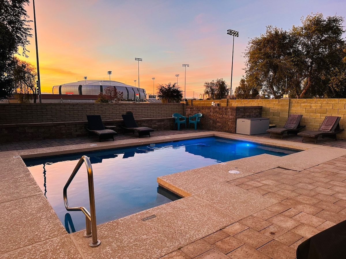 An inviting pool area is highlighted during sunset, with warm colors reflecting on the water's surface. Two lounge chairs are positioned beside the pool, and minimalist outdoor seating is visible in the background. The distant stadium lights add a subtle glow to the scene.