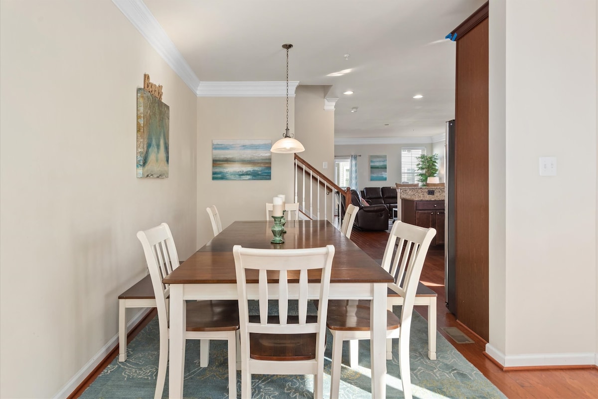 A dining area features a long wooden table surrounded by six white chairs, set against a backdrop of gentle, neutral walls. A pendant light hangs above the table, while two pieces of wall art and an area rug enhance the space's warmth.