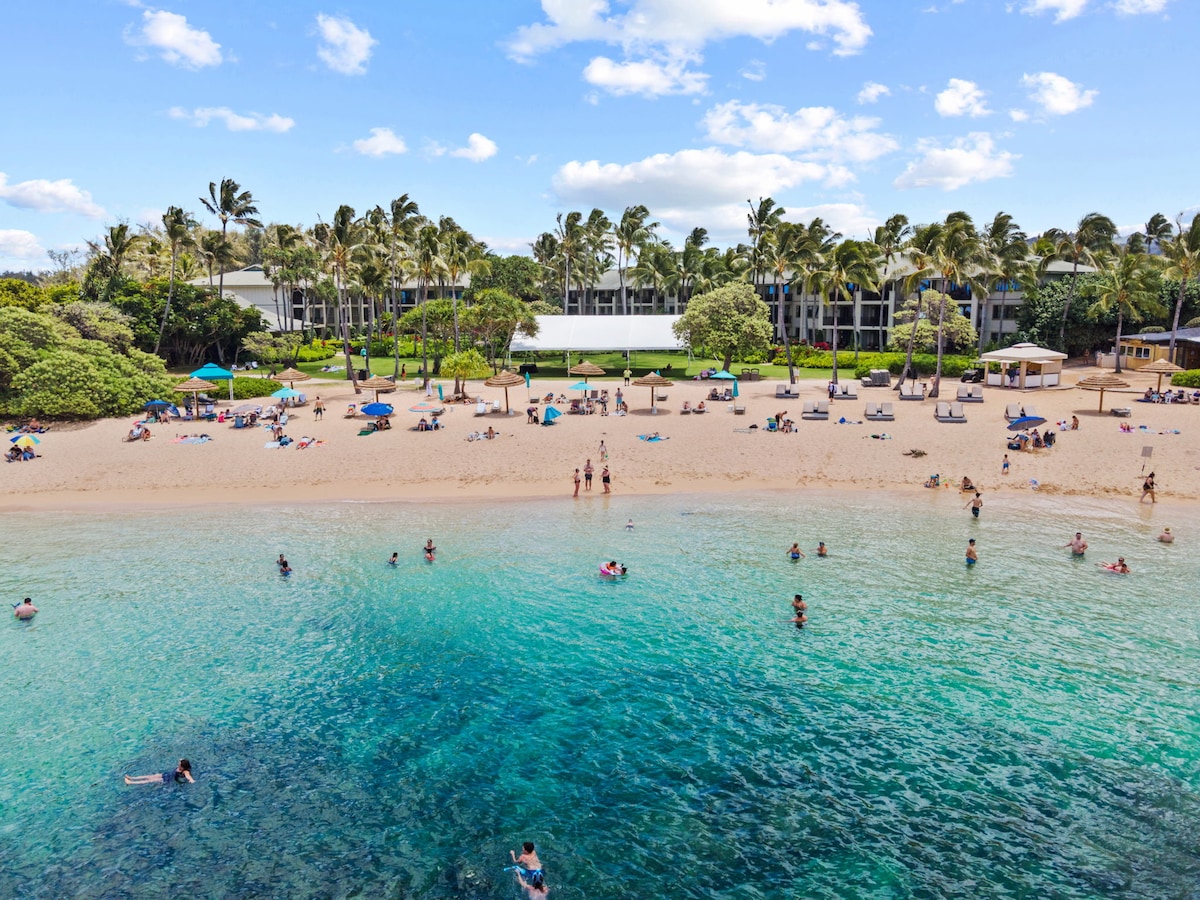 The image captures a scenic beach scene with people enjoying the clear, turquoise water. The shoreline features soft sand and scattered umbrellas for shade. In the background, the condominium units are framed by swaying palm trees, providing a glimpse of the inviting coastal landscape.