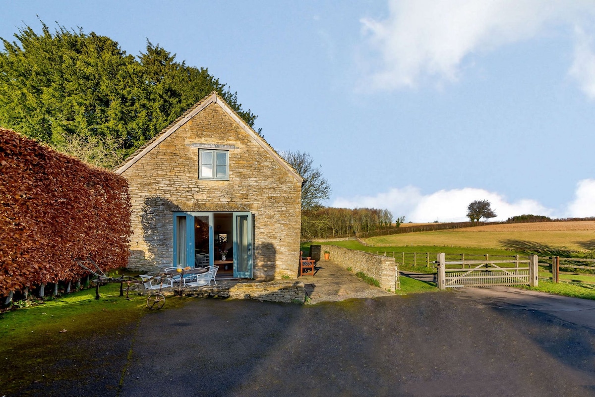 The stone cottage is set against a backdrop of rolling hills and greenery. French doors open to a welcoming outdoor seating area, framed by manicured hedges. The flat driveway leads up to the property, with a gravel path guiding visitors to the main entrance.