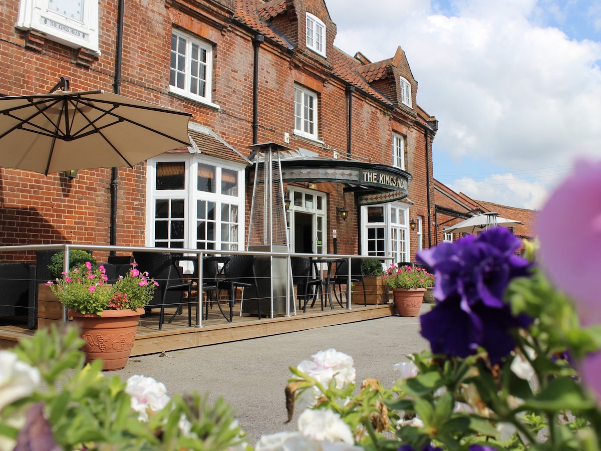 The front terrace of The Kings Head Hotel is shown, featuring several outdoor seating arrangements under a large umbrella. Potted flowering plants in hues of purple and white border the patio, with the hotel's brick exterior and large windows providing a welcoming façade.