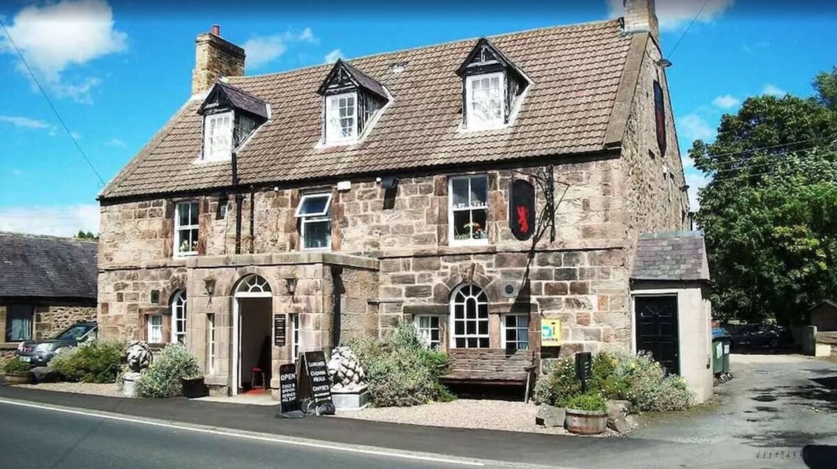 A classic stone building dating back to the late 1700s is featured, showcasing a sloped roof and multiple windows. The exterior is accented by decorative shutters and a welcoming entrance. Stone planters with greenery frame the entryway, with a sign indicating the establishment's name nearby.