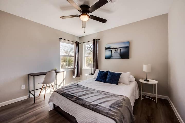 ห้องนอนที่อบอุ่นพร้อมเตียงหรูหราผ้าปูที่นอนสีขาวกรอบและสีเทา โต๊ะข้างเตียงพร้อมโคมไฟที่มีแสงสว่างอบอุ่นช่วยเพิ่มเสน่ห์ในขณะที่โต๊ะและเก้าอี้ที่ทันสมัยสร้างพื้นที่ทำงานที่ใช้งานได้ดี หน้าต่างบานใหญ่นำแสงธรรมชาติเข้ามา