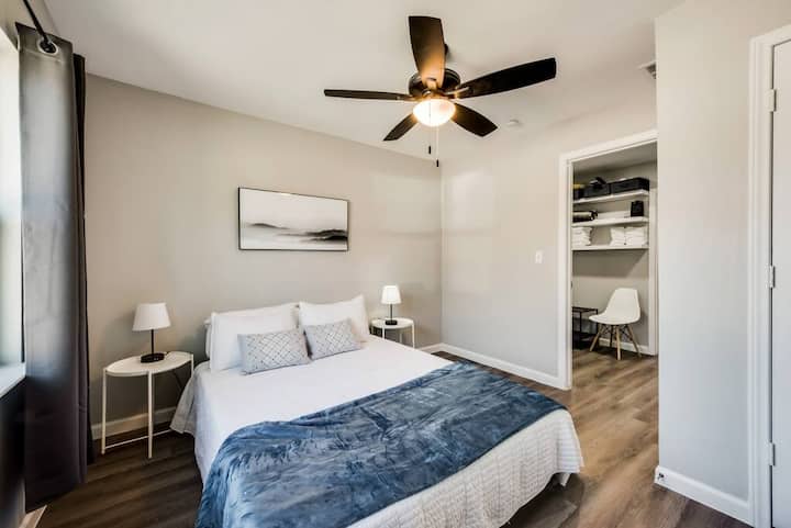 ห้องนอนที่เงียบสงบพร้อมเตียงควีนไซส์ผ้าปูที่นอนสีขาวคมชัดและสีกรมท่า การจับคู่โต๊ะข้างเตียงกับโคมไฟให้ความสมมาตรในขณะที่ตู้เสื้อผ้าแบบเปิดและพัดลมเพดานที่ทันสมัยเพิ่มทั้งสไตล์และฟังก์ชั่น