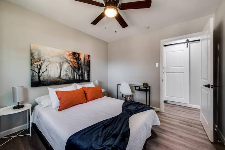 ห้องนอนมีสไตล์พร้อมเตียงหรูหราผ้าปูที่นอนสีขาวคมชัดและหมอนสีส้มเข้ม การขว้างปาสีเข้มช่วยเพิ่มความแตกต่างในขณะที่ภาพวาดภูมิทัศน์โต๊ะข้างเตียงพร้อมโคมไฟและโต๊ะทำงานที่โฉบเฉี่ยวสร้างความรู้สึกอบอุ่นและทันสมัย