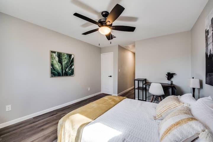 ห้องนอนสไตล์โมเดิร์นที่มีเตียงหรูหราพร้อมผ้าปูที่นอนสีขาวและผ้าปูที่นอนสีทอง โต๊ะทำงานและเก้าอี้สีดำทันสมัยช่วยเพิ่มฟังก์ชันการใช้งานในขณะที่งานศิลปะที่เต็มไปด้วยใบไม้และแสงไฟที่อบอุ่นสร้างบรรยากาศที่อบอุ่นและมีสไตล์