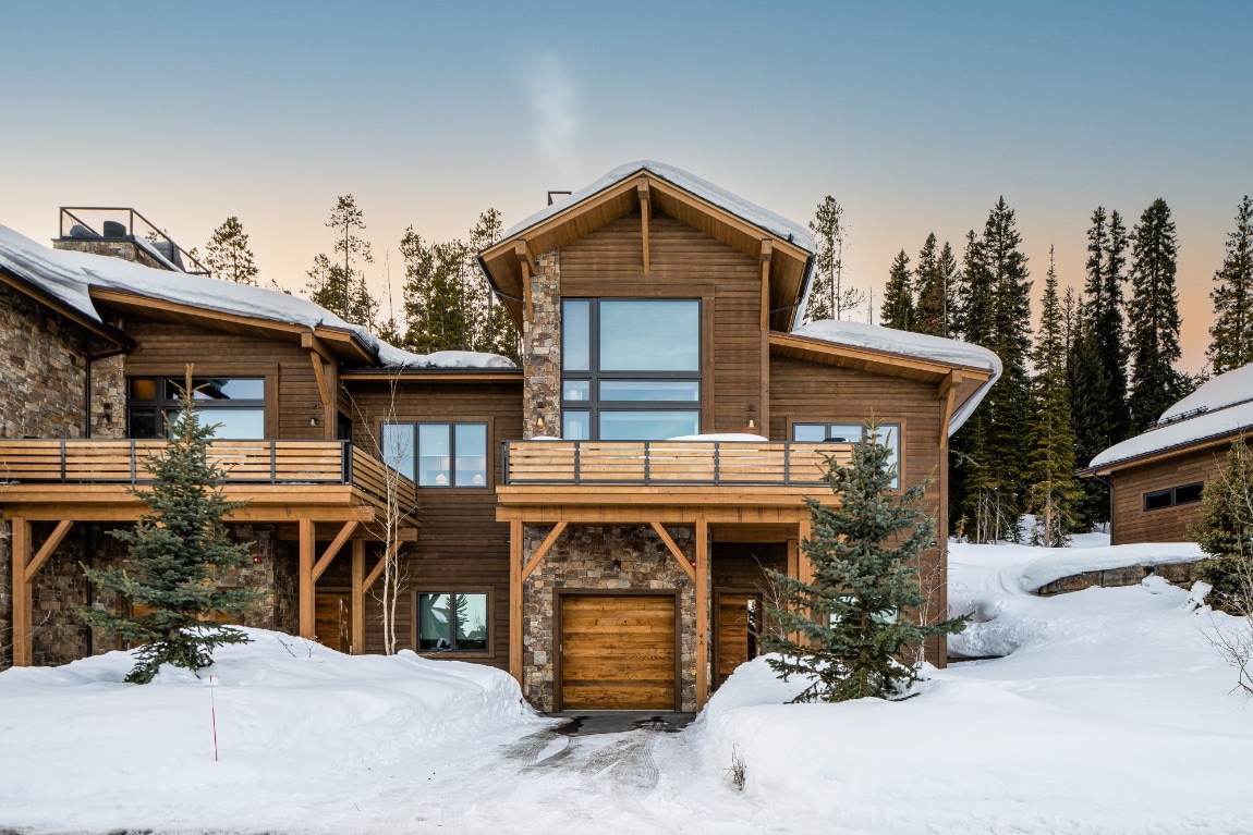 A modern townhome is nestled in a serene snowy landscape, featuring wooden and stone accents. Large windows allow for ample natural light, while trees provide a backdrop. A smooth driveway leads to a garage, accommodating winter access.