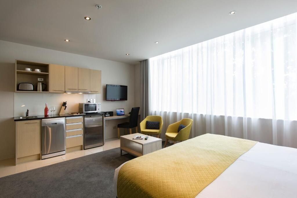 A studio apartment showcases a fully equipped kitchenette with modern appliances, including a refrigerator and microwave. Two cozy armchairs are positioned beside a large window draped in sheer curtains, allowing natural light to fill the space. A spacious bed is centered in the room, offering comfort.