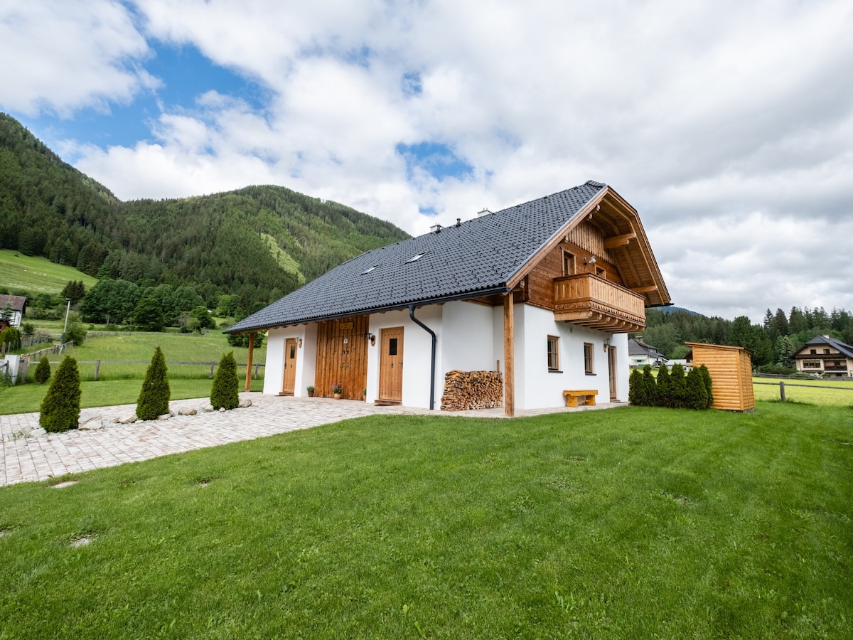A charming chalet is set against a backdrop of green mountains, featuring a sloped roof and wooden accents. The exterior showcases a spacious yard with grass, brick pathways, and neatly trimmed shrubs. A stacked woodpile adds to the rustic atmosphere, while a small storage shed is visible.