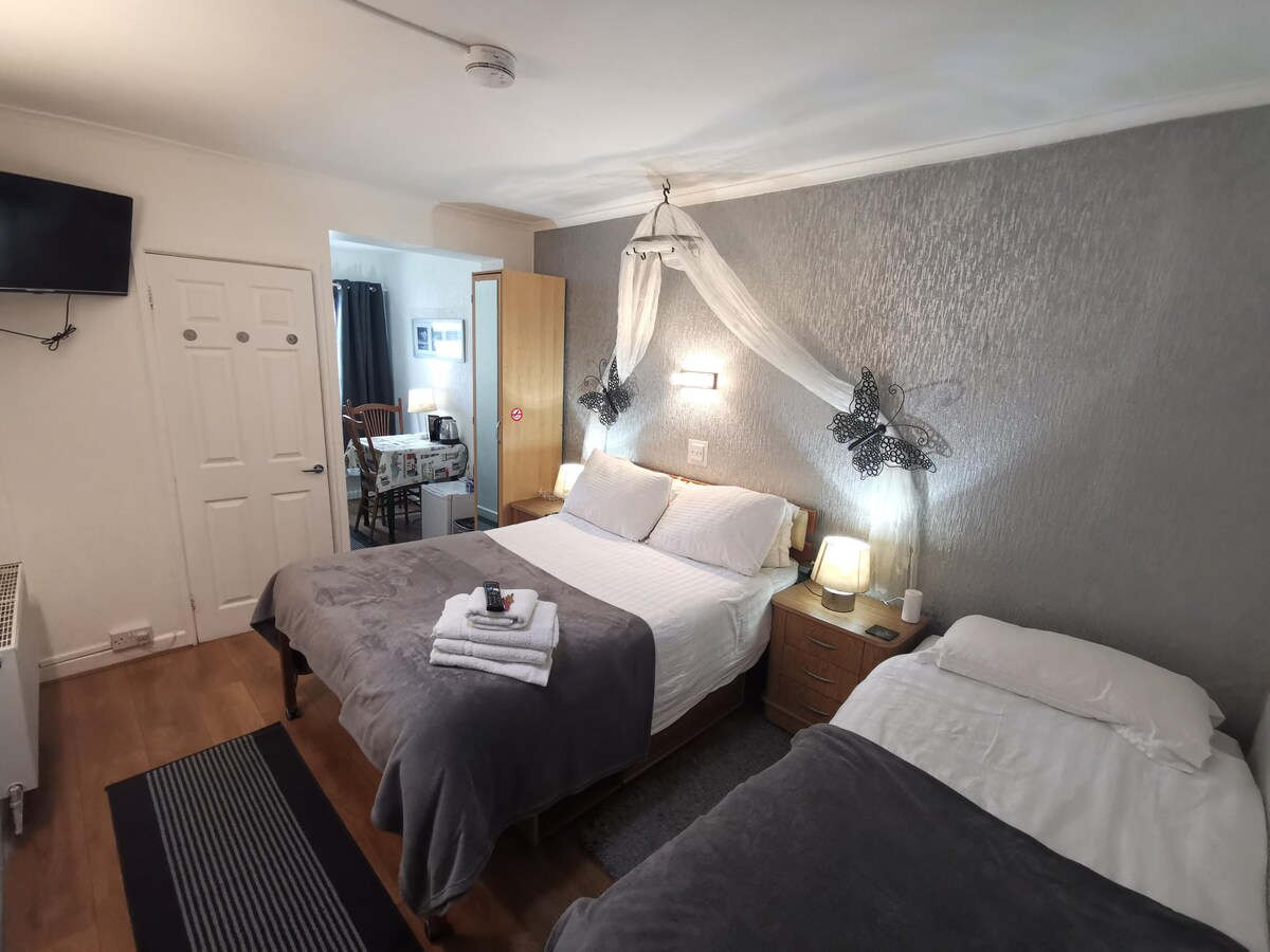 A comfortable bedroom is presented with two spacious beds, each topped with neatly arranged linens and towels. Soft lighting is provided by bedside lamps. A wall-mounted TV is visible, along with a door leading to an adjoining space, enhancing the room's functionality.