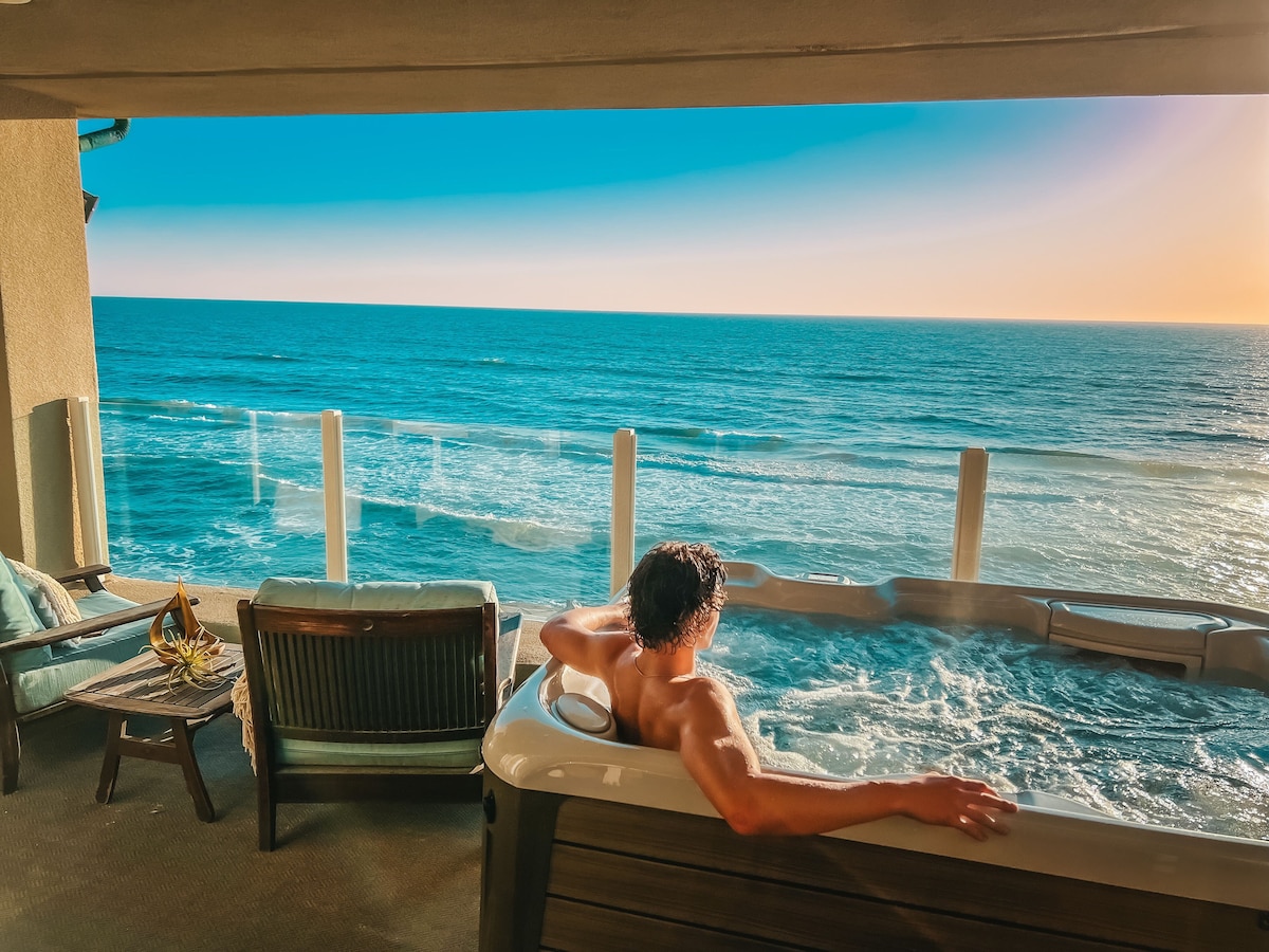 A private spa overlooks the ocean, with a person relaxing in the water. Comfortable seating is arranged nearby, set against a stunning view of the waves and a clear blue sky. Sunlight casts a warm glow over the scene, enhancing the tranquil atmosphere.
