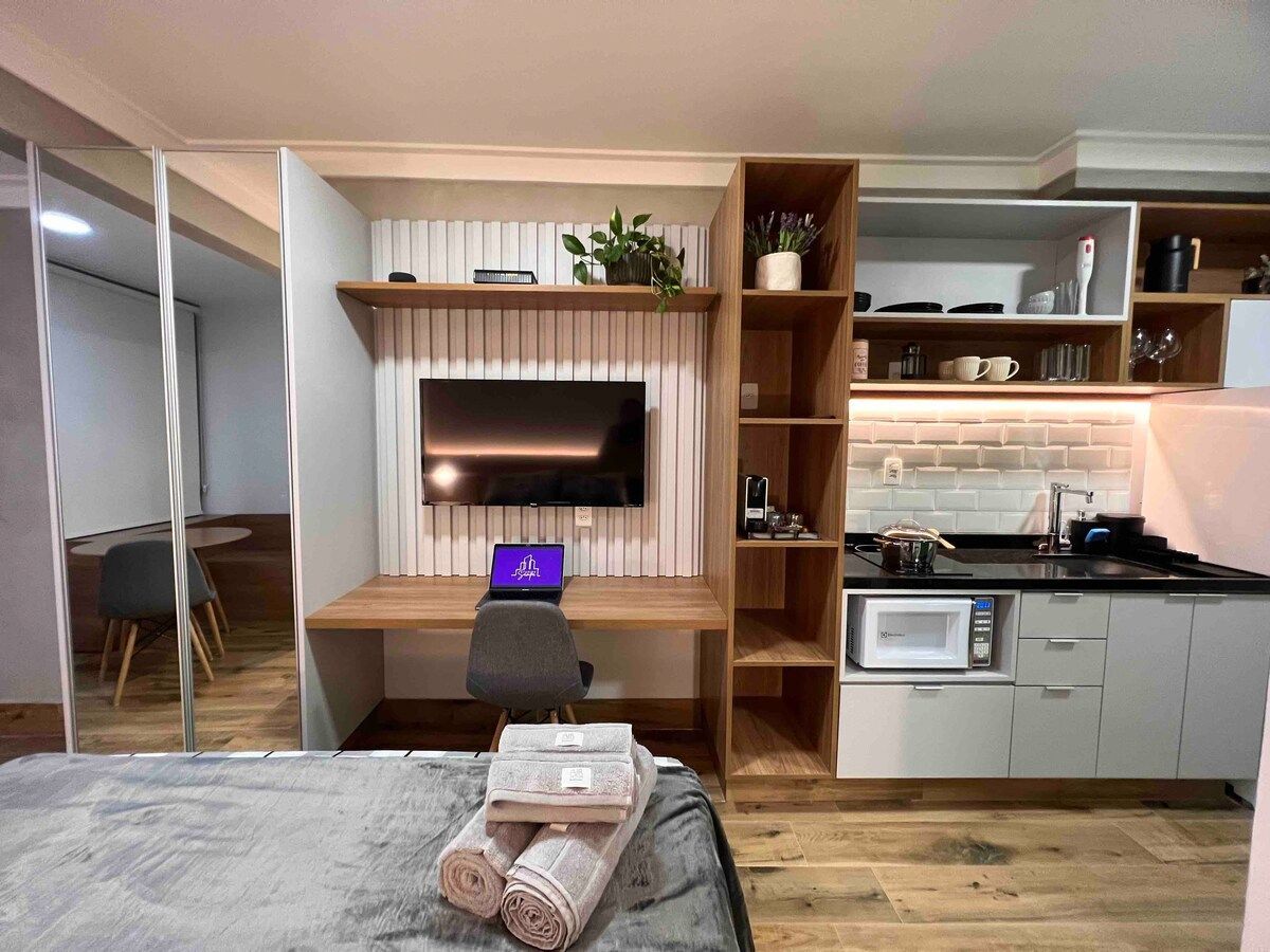 A modern studio unit features a compact kitchen area equipped with a microwave and essential cookware. A workspace with a monitor is adjacent to the entertainment unit, which houses a flat-screen TV. Shelving displays decorative plants and kitchenware, contributing to a functional layout.