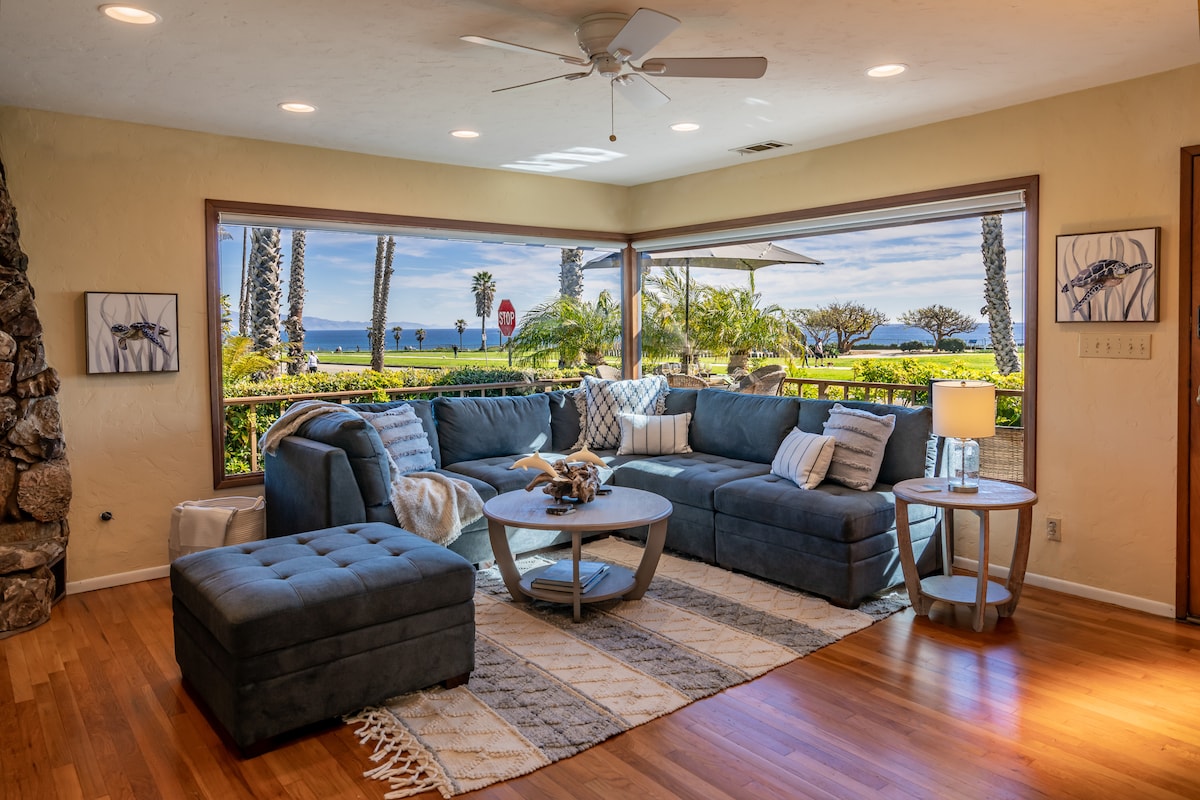 The living room features a spacious sectional sofa arranged around a circular coffee table, with large bay windows offering views of the ocean and surrounding greenery. A textured rug lies beneath, and a ceiling fan is mounted above for comfort. Natural light fills the space.