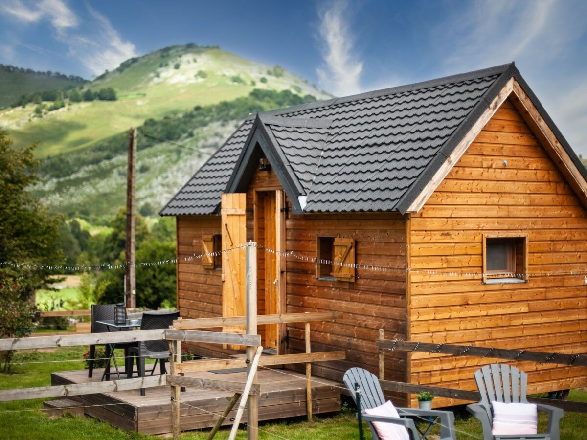 A wooden cabin is set against a backdrop of rolling green hills under a partly cloudy sky. The structure features a peaked roof, a central entrance, and several windows. Outdoor seating areas with tables and chairs provide space to relax in the surrounding nature.