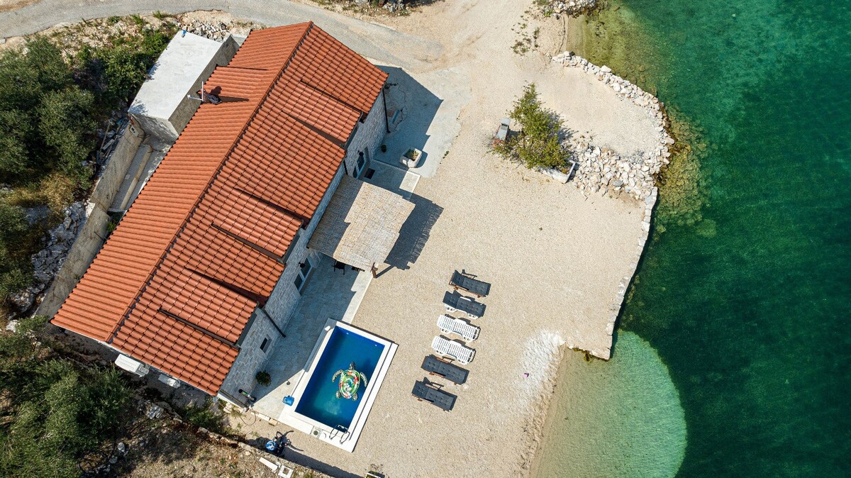 An aerial view showcases the beachfront villa with a red-tiled roof and a private pool. Sun loungers are arranged on the sandy shore, leading to the clear turquoise waters. The surrounding landscape features rocky outcrops and greenery, enhancing the coastal setting.