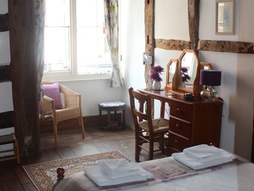 A cozy accommodation is depicted, featuring a comfortable double bed with neatly arranged linens. A wooden desk with a mirror and a lamp is set beside a wicker chair. Natural light enters from a large window, illuminating the rustic timber beams along the walls.