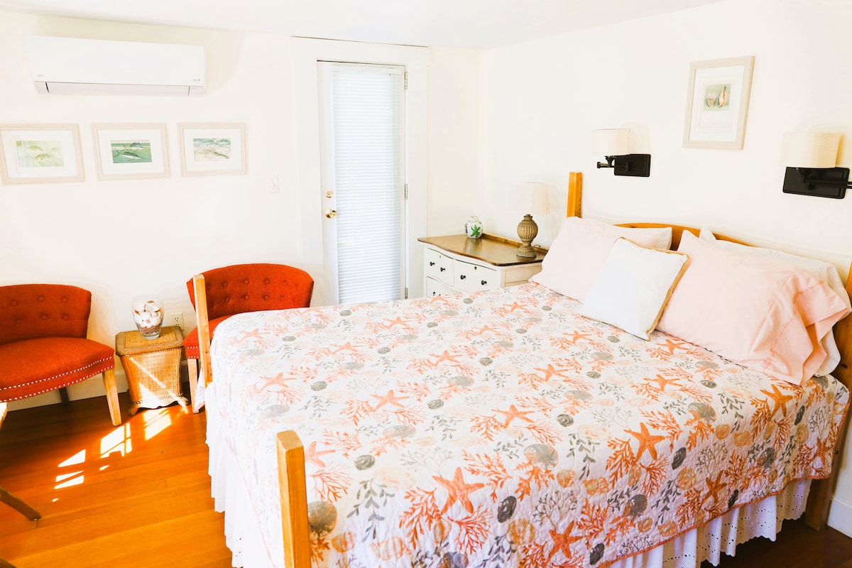 A cozy room features a queen-sized bed adorned with a floral bedspread. A small table with two armchairs is positioned beside the bed, creating an inviting nook. Light flows through a door leading to a private balcony, complemented by soft wall-mounted sconces.