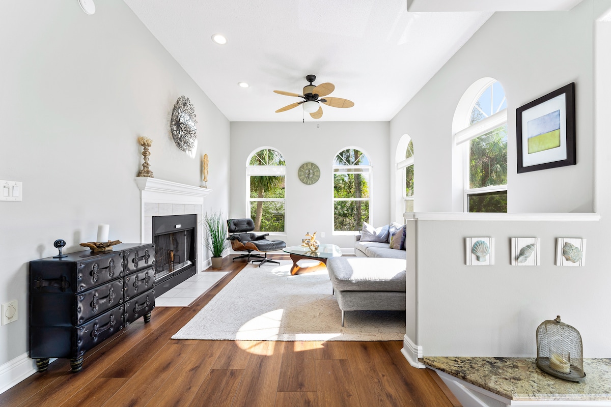 A bright and airy living area features tall arched windows allowing natural light to fill the space. A comfortable gray sectional sofa is arranged around a central area rug, complemented by a stylish black cabinet and an elegant ceiling fan. Decorative elements enhance the inviting atmosphere.
