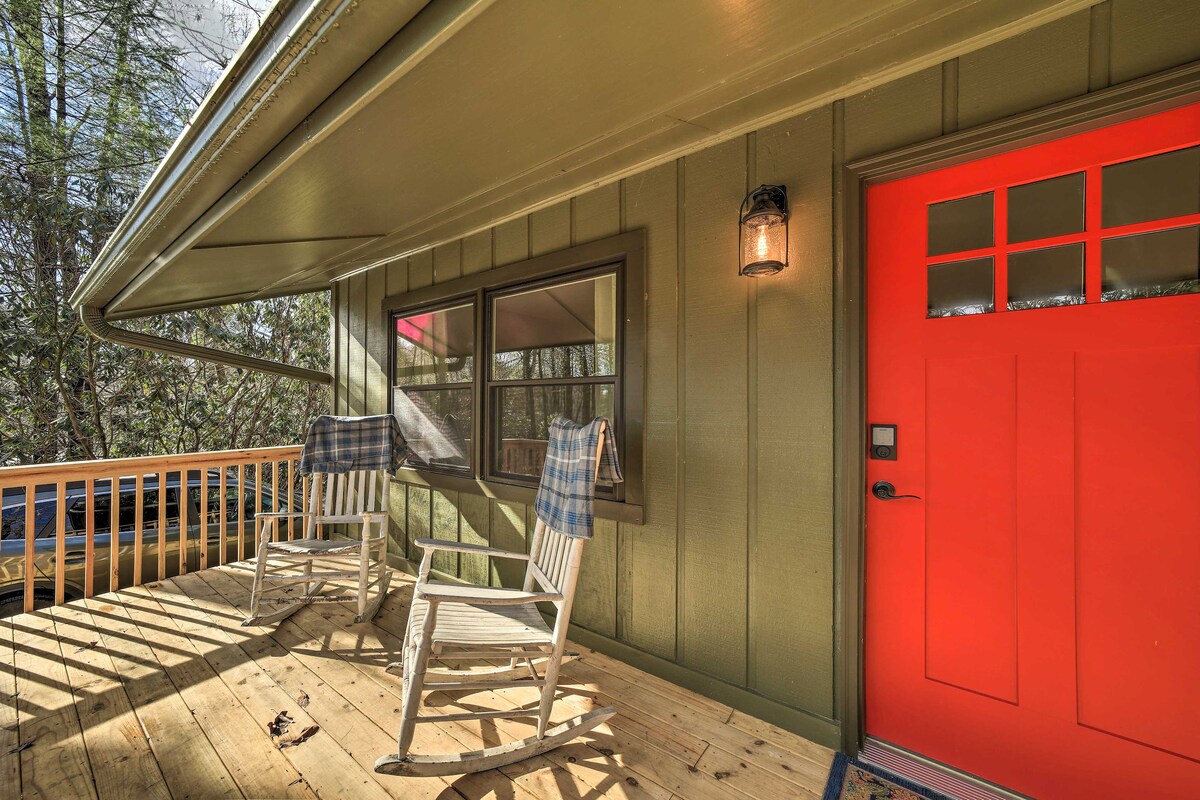 A welcoming front porch features two rocking chairs facing a serene wooded view. The vibrant red door adds a cheerful touch, while the wooden floorboards reflect natural light filtering through the surrounding trees.