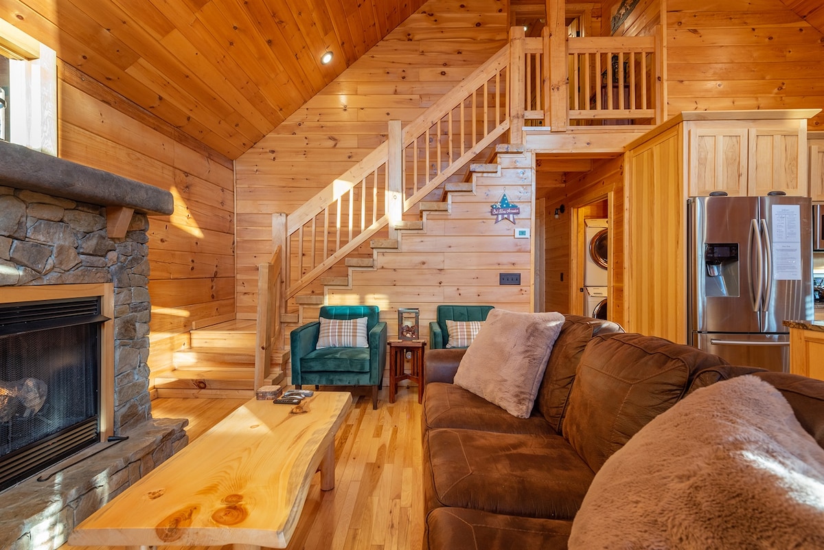 A cozy living space is accentuated by natural wood paneling and a stone gas fireplace. A plush sofa is positioned in front of the fireplace, accompanied by a wooden coffee table. A stairway leads to the upper floor, with two green armchairs in the corner.