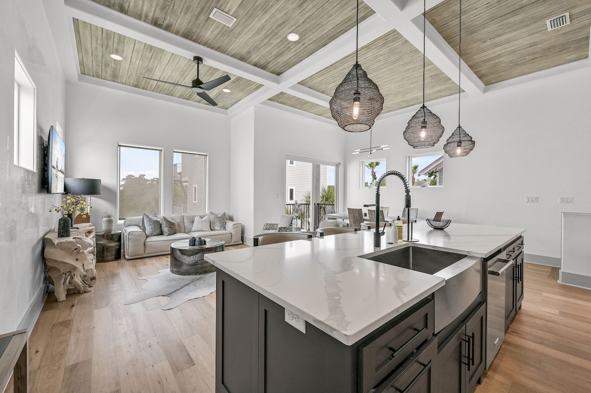 An open-concept living area features a comfortable sectional sofa and sleek coffee table, complemented by large windows allowing natural light. The spacious kitchen island, equipped with modern fixtures and pendant lighting, faces the common area, creating an inviting space for socializing.