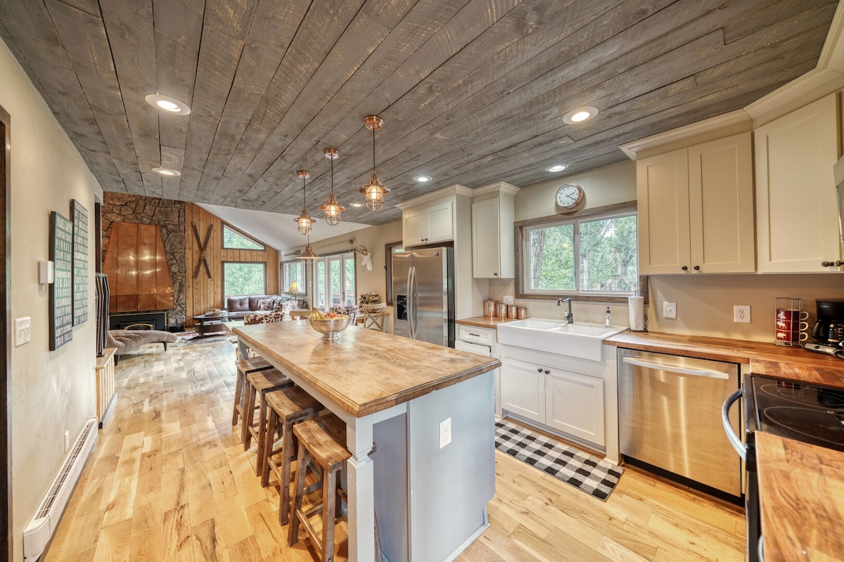 A bright and open kitchen area features a large wooden island with seating and modern appliances. Natural light floods the space through multiple windows, highlighting the wood-textured ceiling and floors. The living area is visible in the background, creating a seamless transition between spaces.