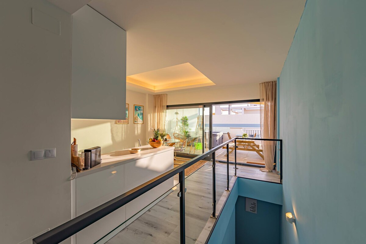 A bright and modern open-plan living area is visible from the upper level. A spacious kitchen is blended with a lounge area, leading to large glass doors that open to the terrace. Soft furnishings and greenery can be seen beyond the doors.
