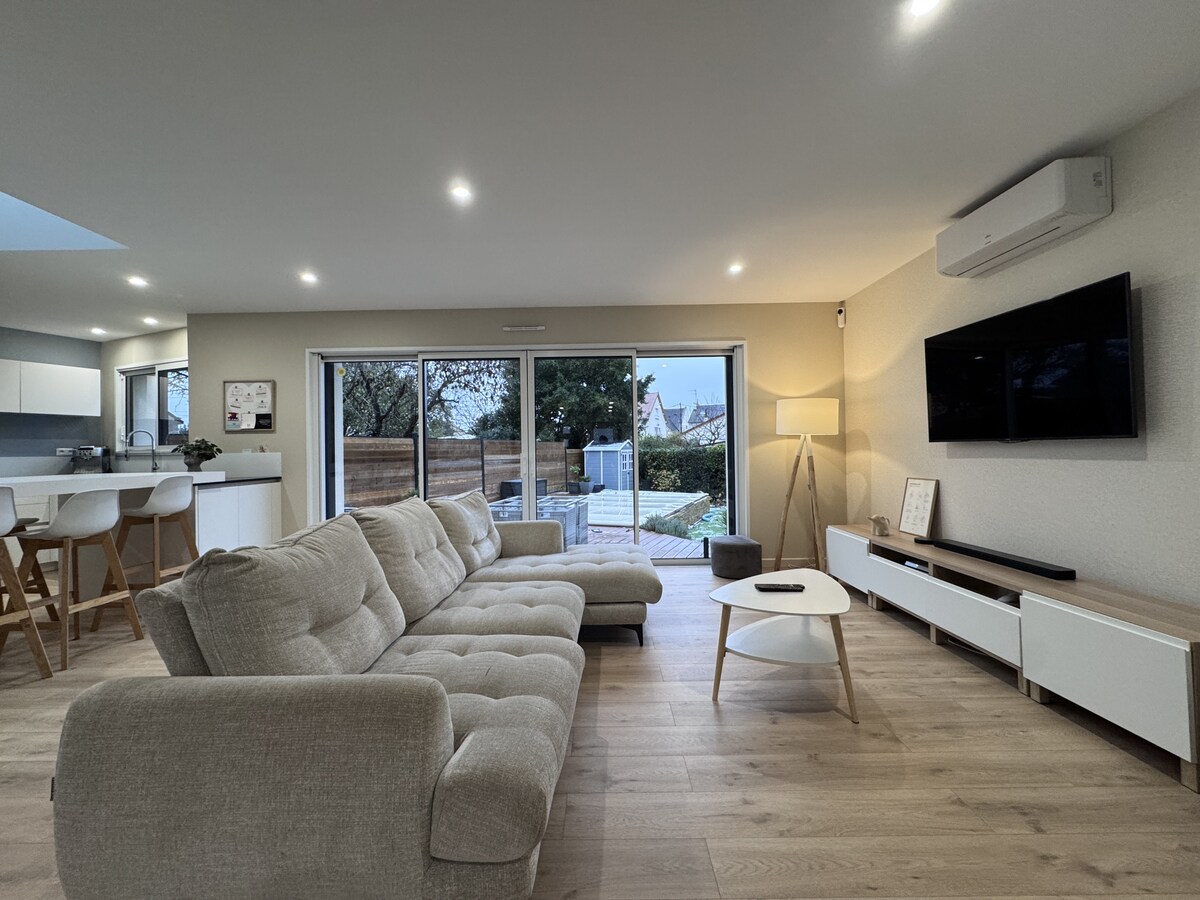 A spacious living area features a light-colored sectional sofa, positioned to face a mounted television. Large glass doors allow for natural light and provide views of the outdoor space. Soft lighting and wooden flooring enhance the ambiance, complemented by modern decor.