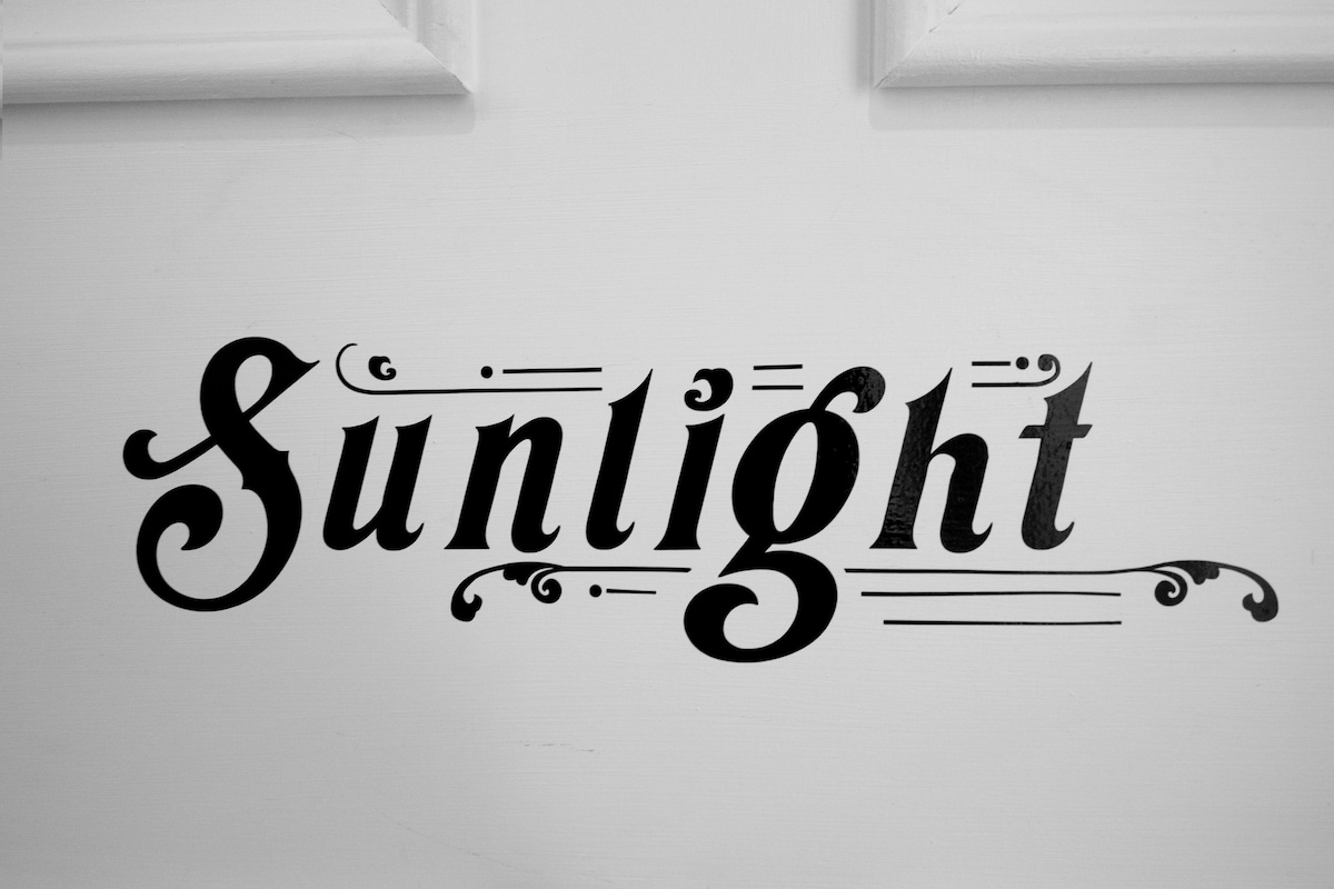 The word 'Sunlight' is displayed in elegant, stylized lettering on a white surface, framed by light-colored borders. The design features decorative flourishes, creating a classic yet contemporary aesthetic.