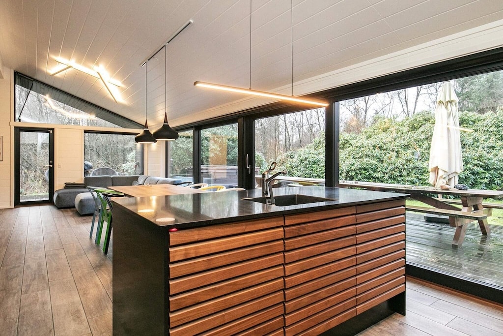 An open kitchen features a contemporary design with a dark countertop and wooden cabinetry. Pendant lights hang above the island, while large glass doors provide a view of the outdoor area. A comfortable seating area is visible, surrounded by greenery.