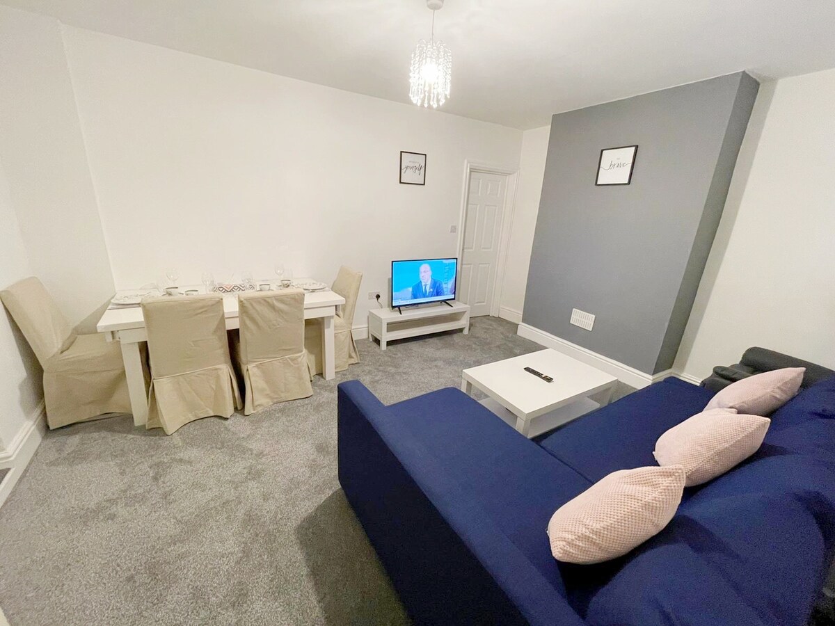 The living area features a comfortable blue sofa with decorative pillows and a white coffee table. A dining area with a light-colored table and matching chairs is visible. A Smart TV is positioned on a low stand, and the space is illuminated by a modern chandelier.