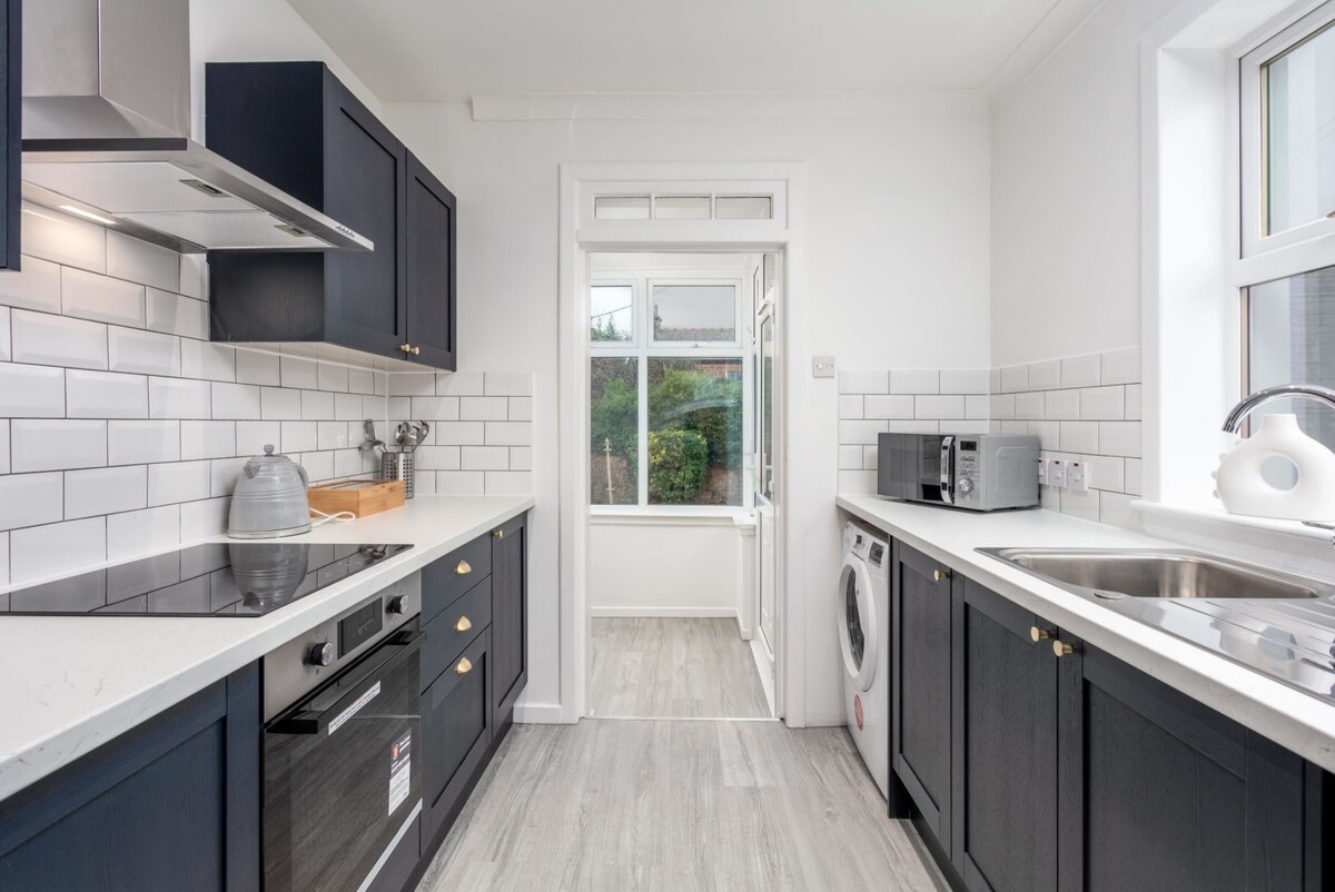 The kitchen features dark cabinetry and sleek countertops, complemented by white tiled backsplash. A modern oven and stovetop are present, along with a microwave and washing machine. A large window brings in natural light, enhancing the space, while a light-colored floor adds a contemporary touch.