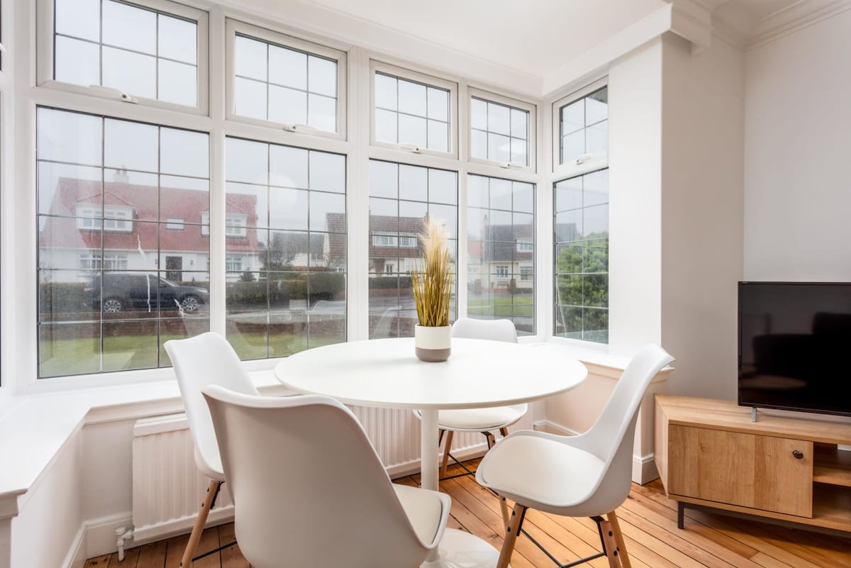 A bright dining area is featured with a round table surrounded by four white chairs. Large windows provide views of the outside while allowing natural light to illuminate the space. A wooden media console sits nearby, enhancing the modern aesthetic.