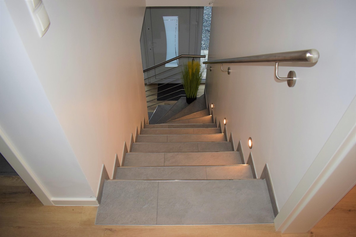 A staircase equipped with contemporary lighting guides the way downwards. The walls are finished in a light tone, complemented by sleek stone tiles underfoot. A decorative plant is positioned at the bottom of the stairs, contributing to the clean and modern aesthetic of the space.