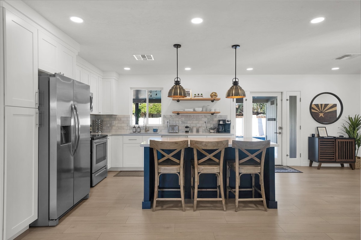 The kitchen features stainless steel appliances and ample counter space. A large island with seating for four is illuminated by pendant lights. White cabinetry and a tile backsplash enhance the modern look, while natural light fills the room through windows overlooking the backyard.